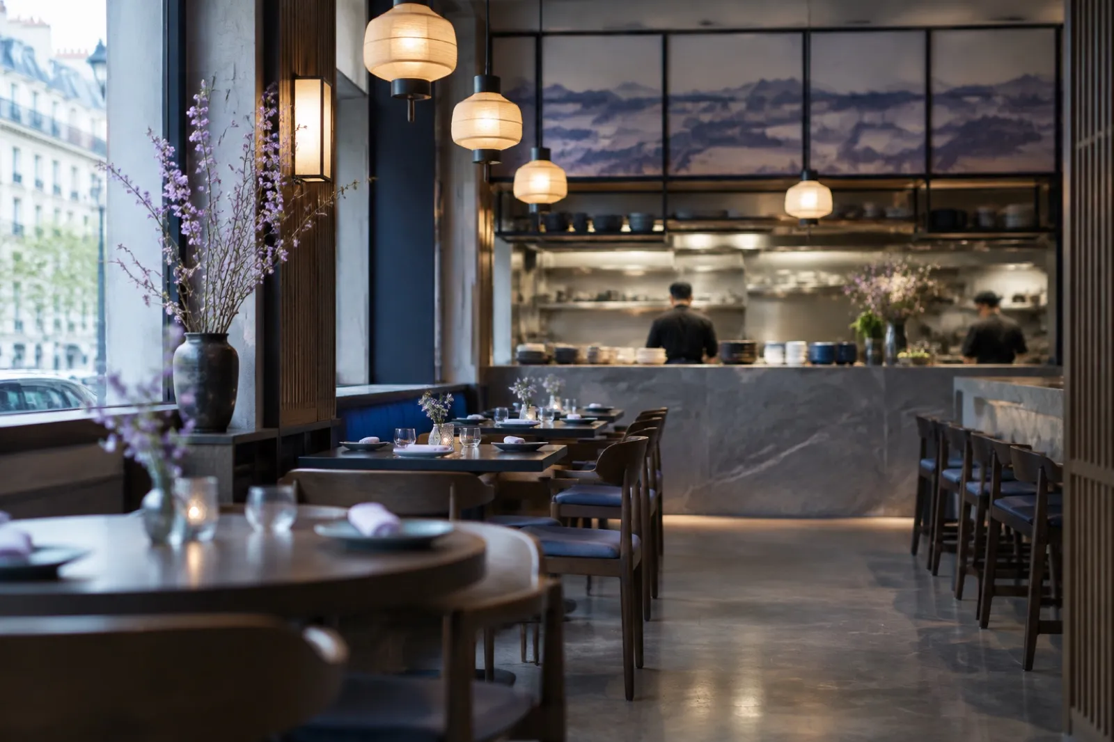Salle de restaurant avec cuisine ouverte, tables dressées et suspensions allumées